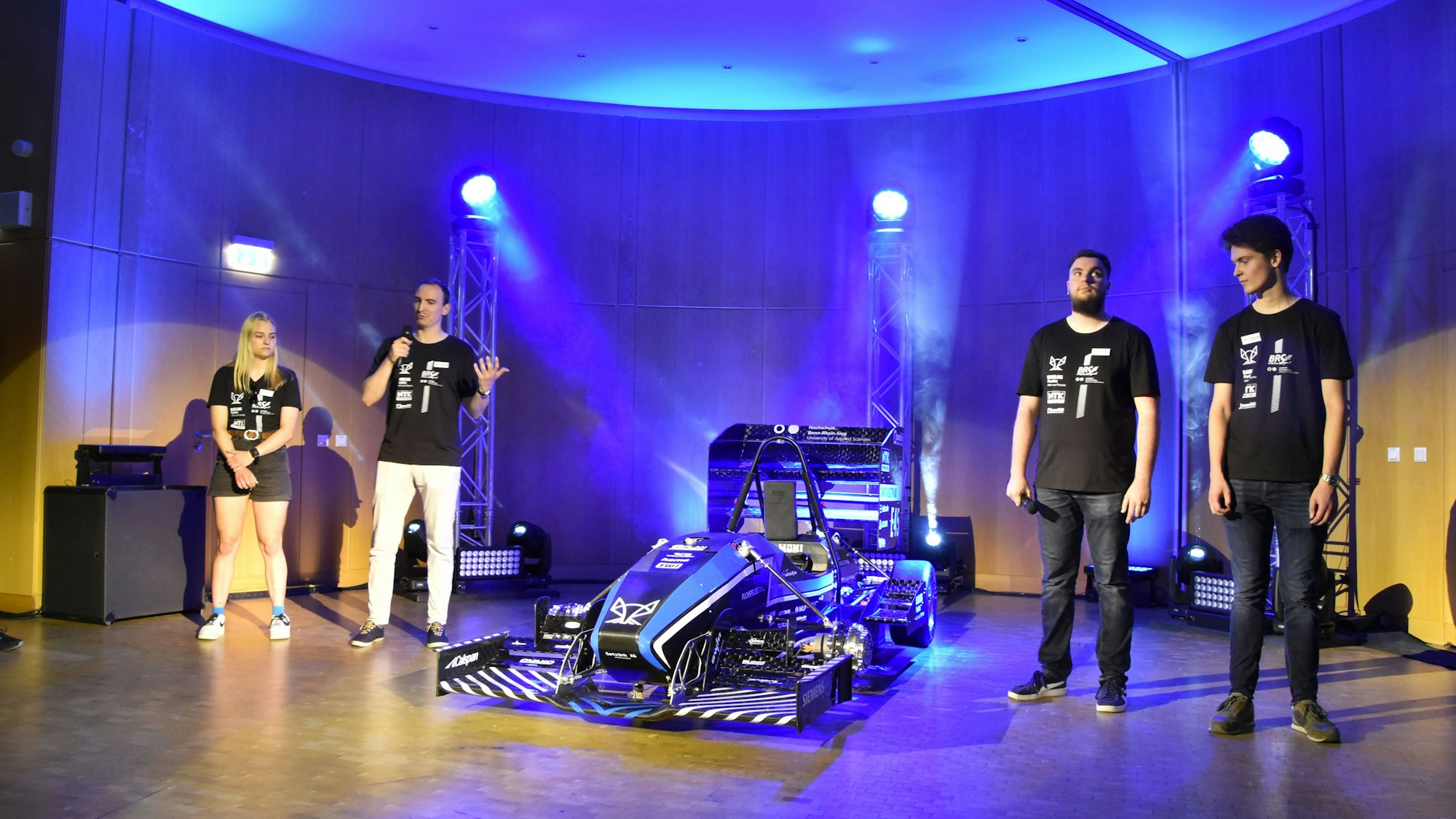 Rollout des neuen Rennwagens der Hochschule Bonn-Rhein-Sieg: Experten erklären die neu entwickelten Bauteile. Drei Männer und eine Frau stehen neben dem Rennwagen auf der Bühne.