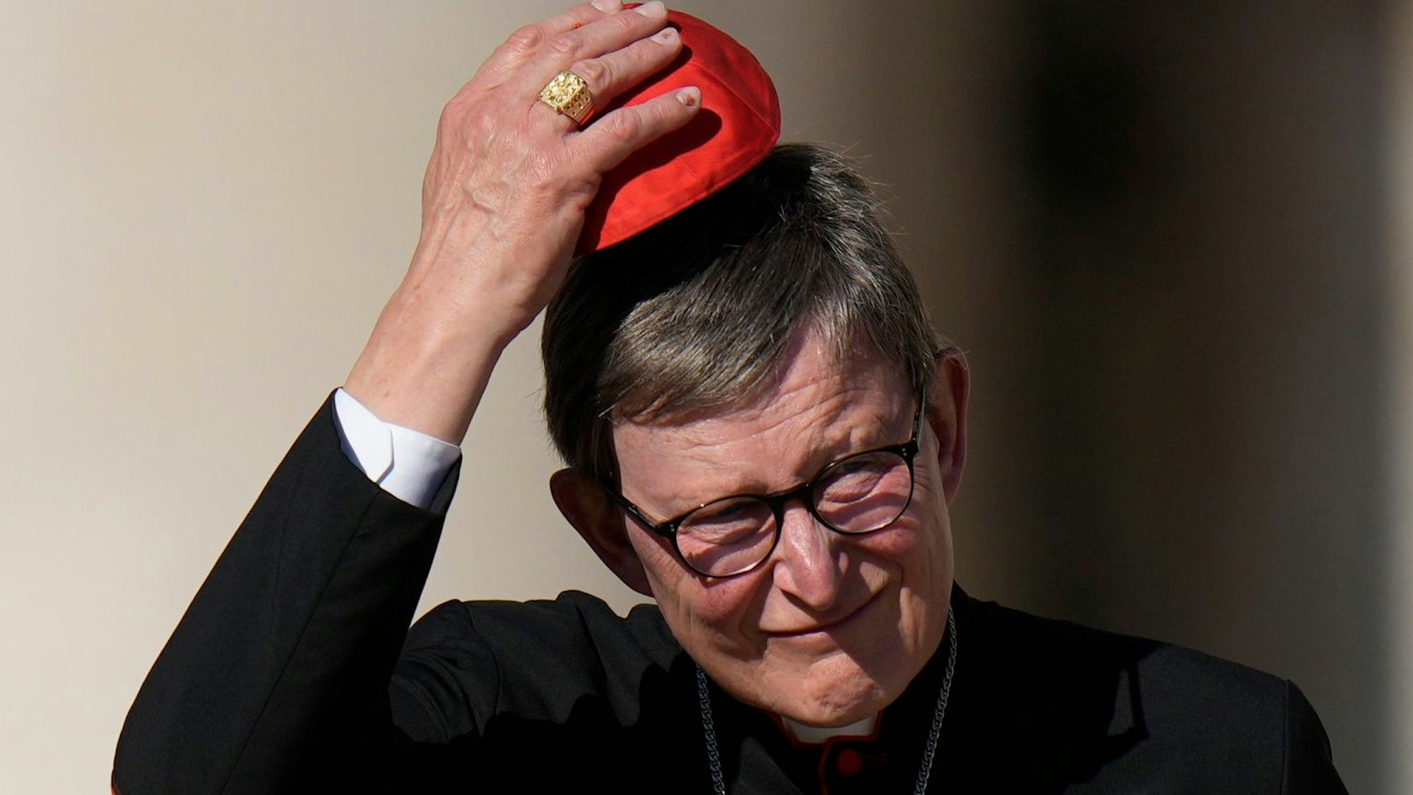 Kardinal Rainer Maria Woelki in schwarzem Talar und einer roten Kappe, dem Pileolus, vor unscharfem Hintergrund. Mit einer Hand hebt er die Kappe in die Höhe. Vor der Brust hängt ein silbernes Kruzifix.