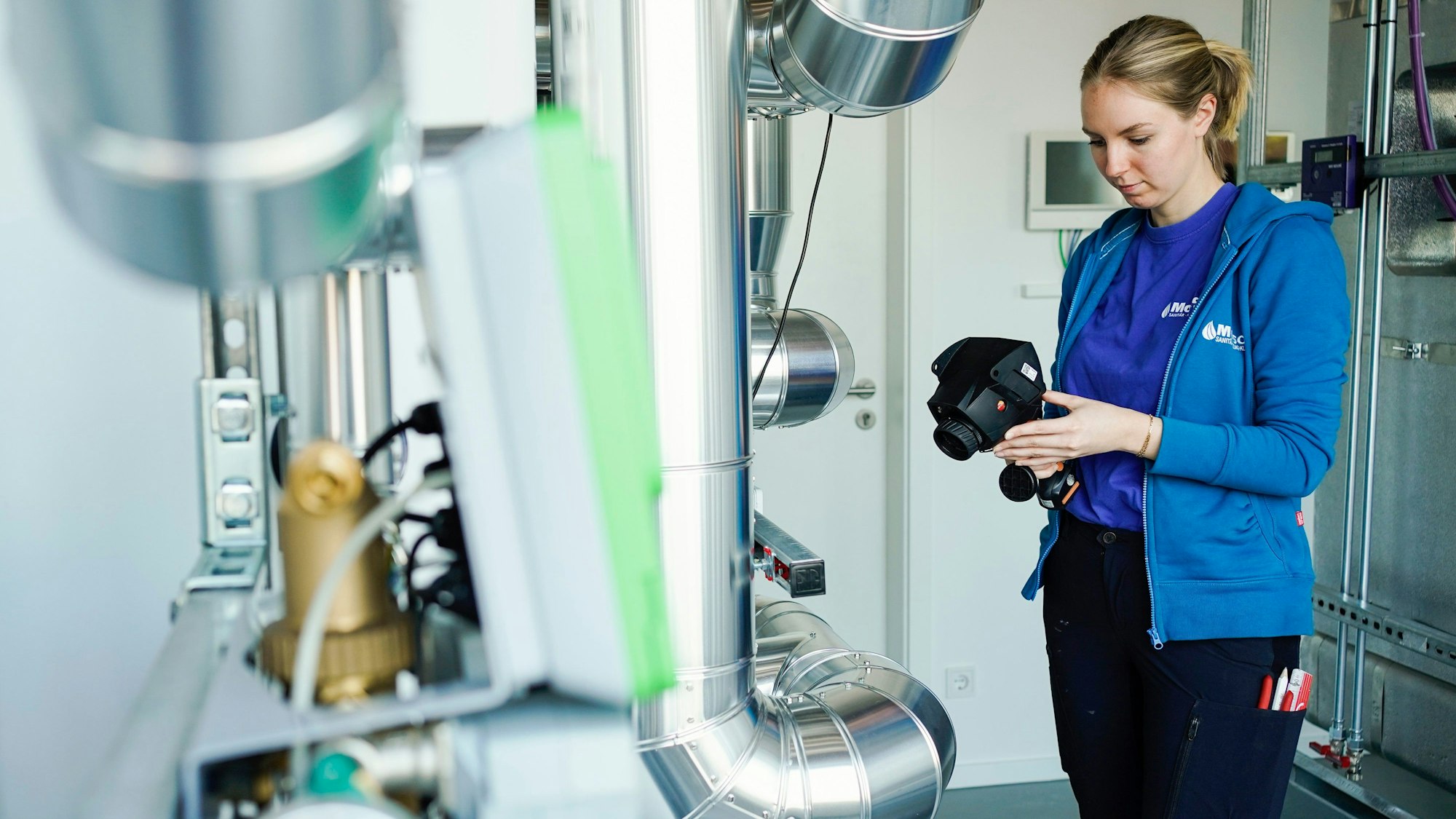 Eine Auszubildende steht in einem Betrieb mit einem Wärmemessgerät an einer Wärmepumpe.