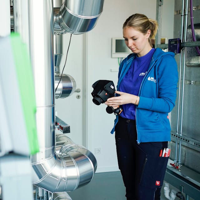 Eine Auszubildende steht in einem Betrieb mit einem Wärmemessgerät an einer Wärmepumpe.