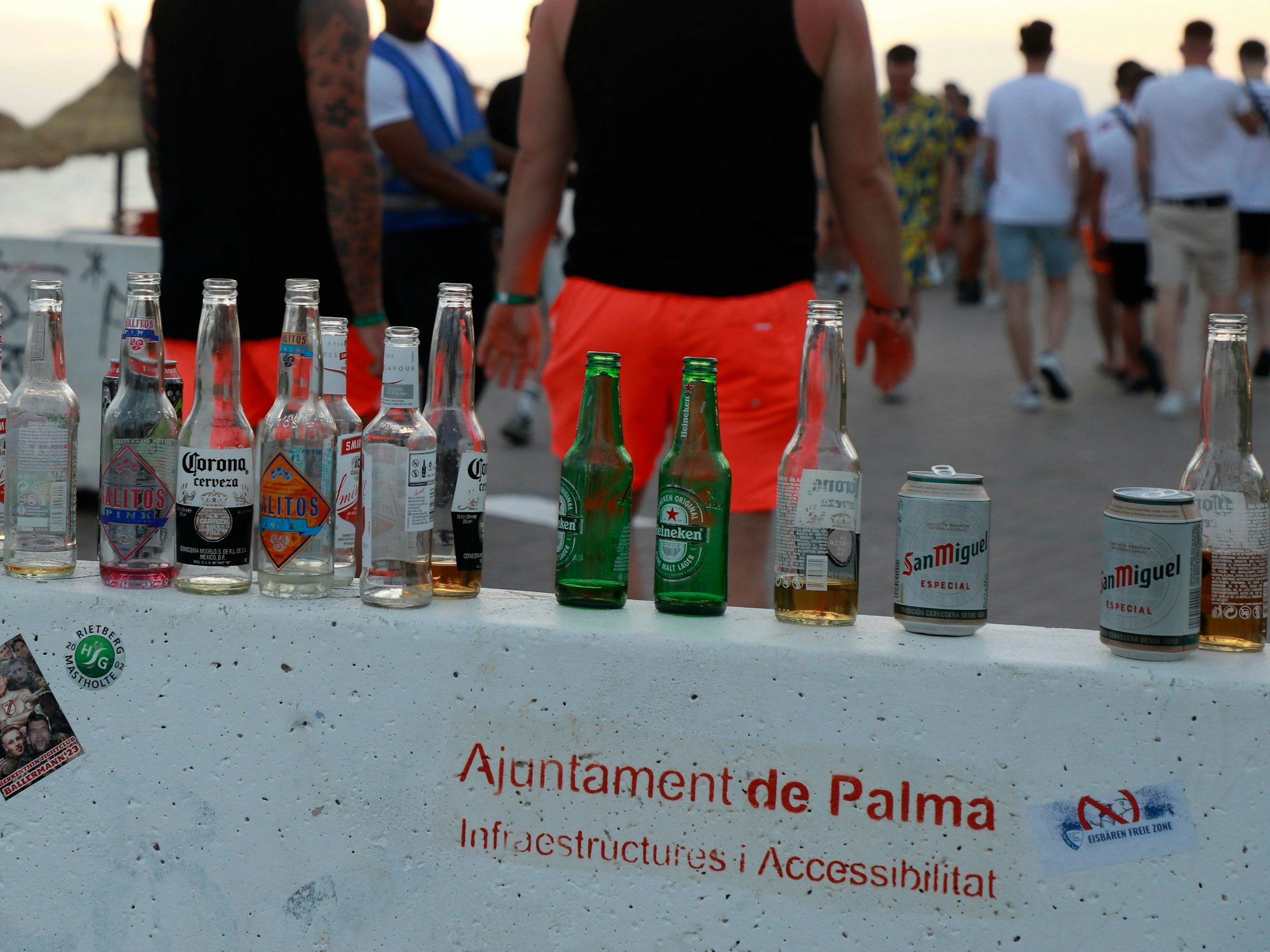 Menschen haben sich am Abend an der Promenade am Strand von Arenal versammelt. Auf einer Mauer stehen mehrere leere Flachen alkoholischer Getränke.