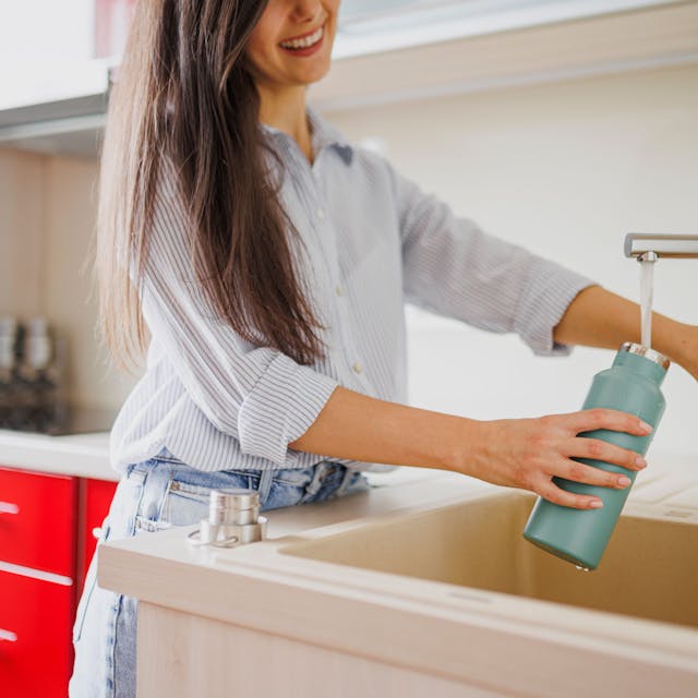 Eine Nahaufnahme einer Frau, die in der Küche eine wiederverwendbare Wasserflasche auffüllt.