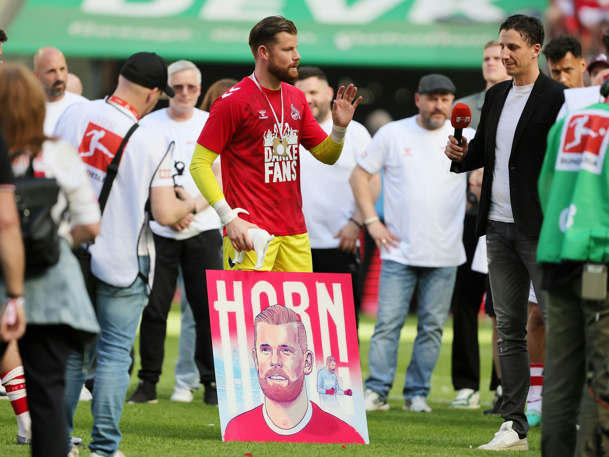 Tim Horn steht auf dem Platz, neben im Christian Keller. Im Hintergrund Steffen Baumgart. Vor Horn steht ein großes Bild, das ihn und seinen Nachnamen zeigt.