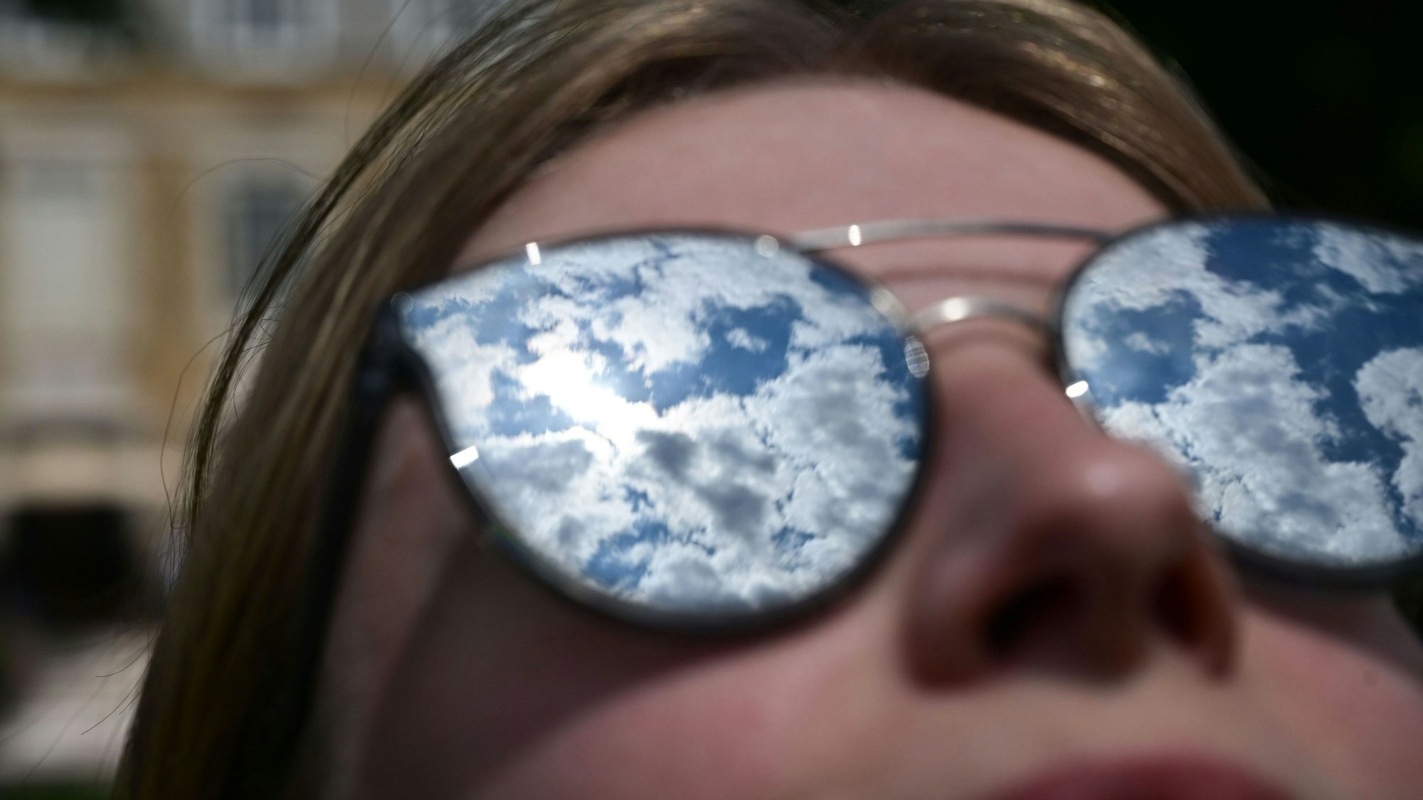 Eine Frau hält ihr Gesicht in die Sonne, die durch leichte Wolken auf ihrer verspiegelten Brille erscheint.