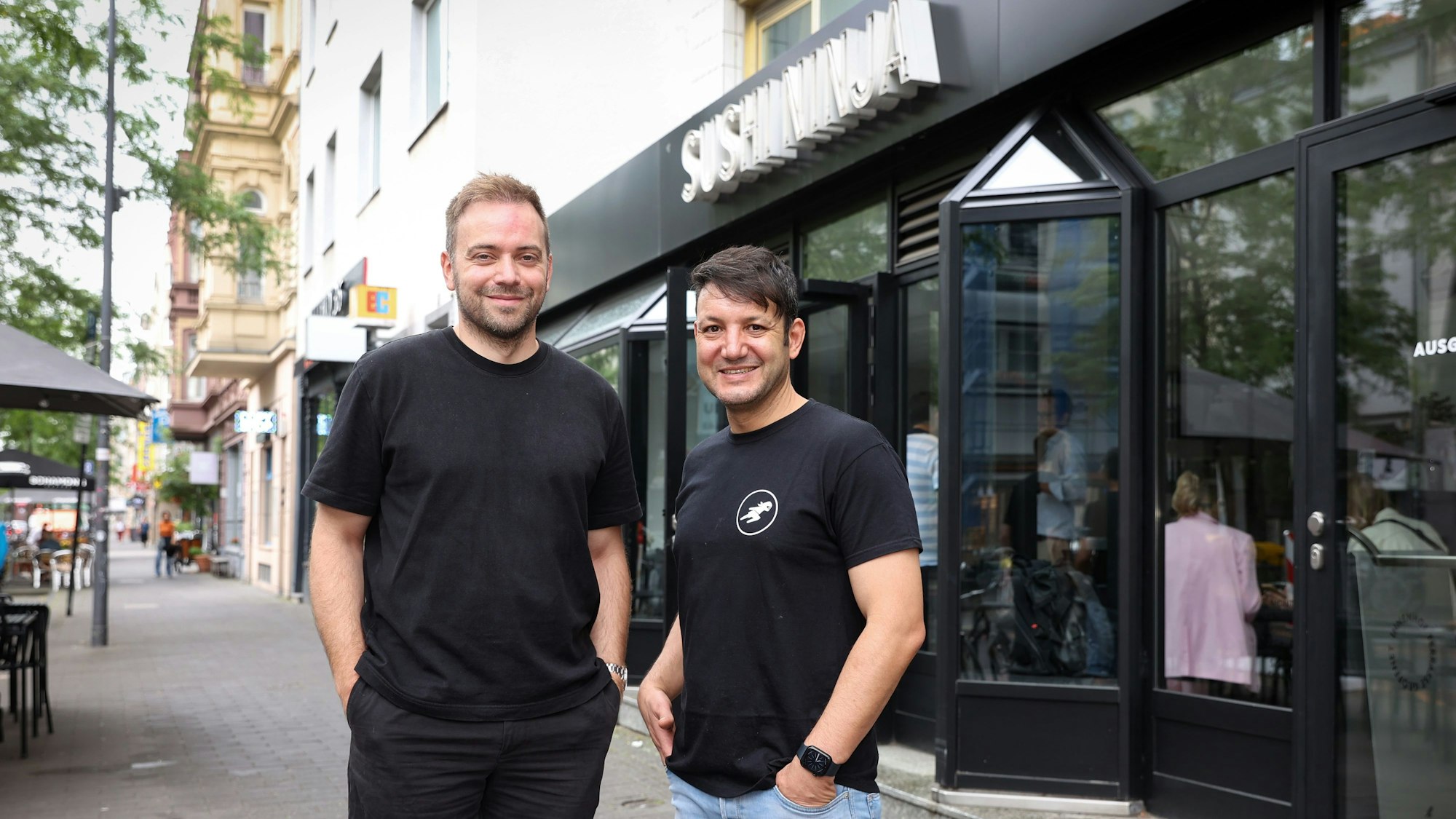 David Keuenhof und Sofien Kouraichi stehen vor einem von drei Kölner Sushi-Ninja-Restaurants. Sie tragen dunkle T-Shirts.