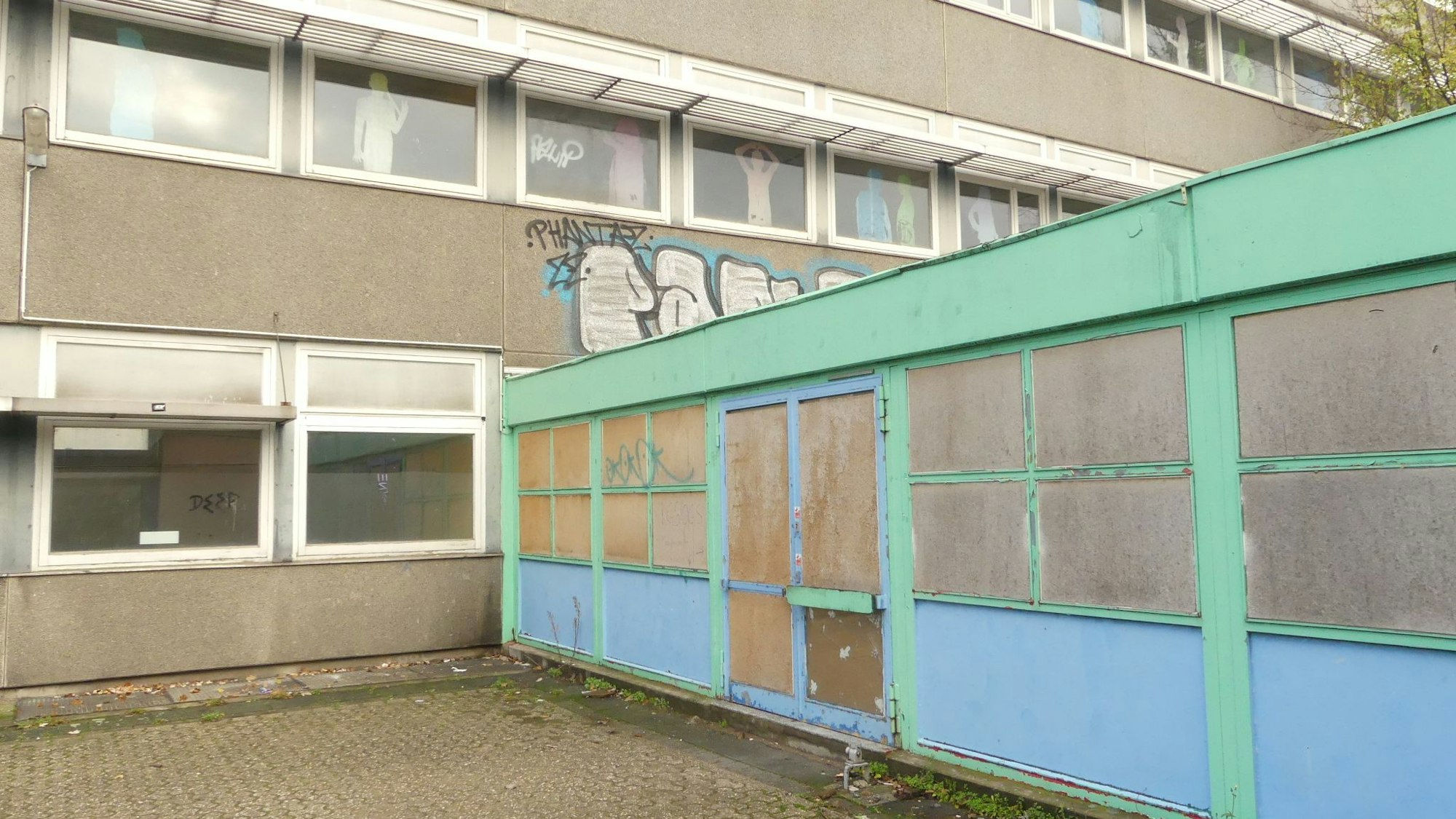 Ein schmuckloses Schulgebäude mit einem grün-blauen Anbau ist zu sehen.