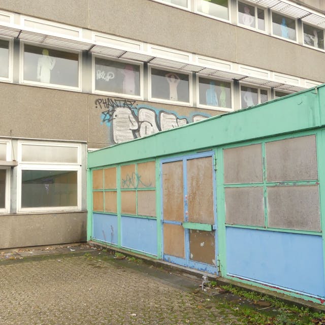 Ein schmuckloses Schulgebäude mit einem grün-blauen Anbau ist zu sehen.&nbsp;