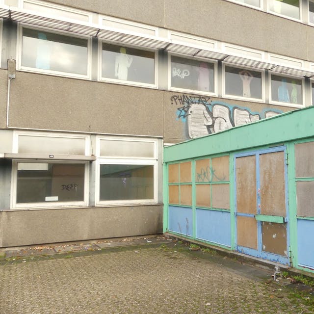 Ein schmuckloses Schulgebäude mit einem grün-blauen Anbau ist zu sehen.&nbsp;