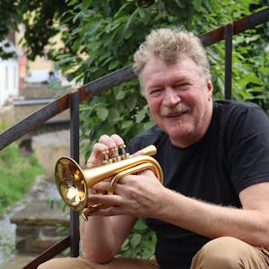 Hans Peter Salentin sitzt im Café Marielle in Bad Münstereifel auf einer rostigen Treppe, in seiner Hand hält er eine Mini-Trompete.