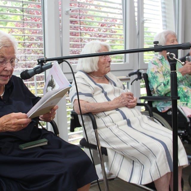 Drei ältere Damen sitzen nebeneinander, eine liest aus einem Buch, sie spricht in ein Mikrofon.