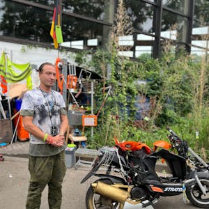 Der Obdachlose Marcel K. steht vor seiner Hütte am Höhenbergbad. Neben ihm ein Motorroller.