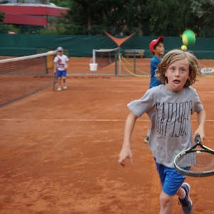 Ein Junge läuft mit einem Schläger über den Tennisplatz. Ein Tennisball fliegt durch die Luft. Der Junge hat einen konzentrierten Gesichtsausdruck.