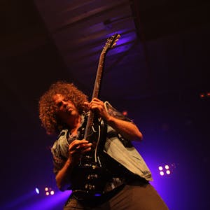 Andrew Stockdale von Wolfmother ist in sein Gitarrenspiel vertieft. Im Hintergrund glimmen die Bühnenscheinwerfer.