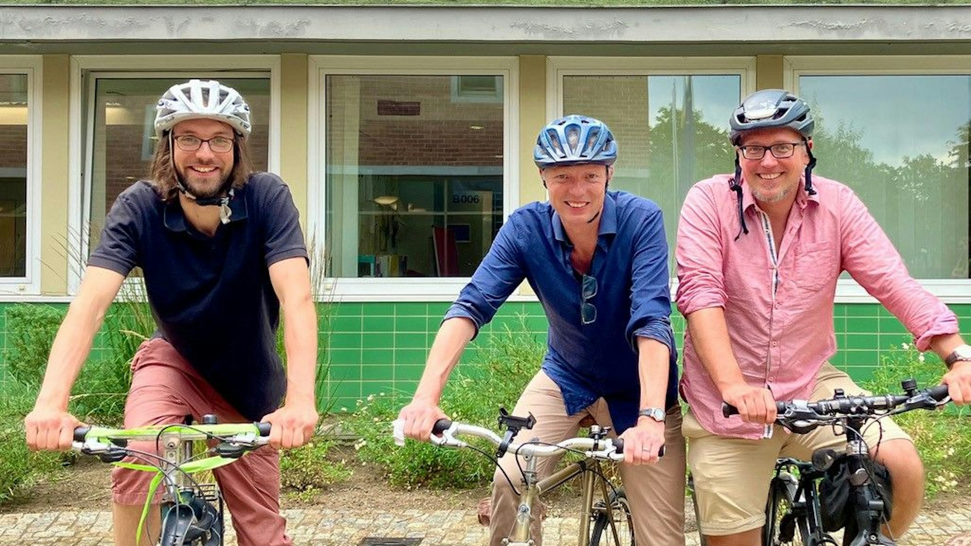 Drei Männer auf Fahrrädern stehen vor einem Schulgebäude