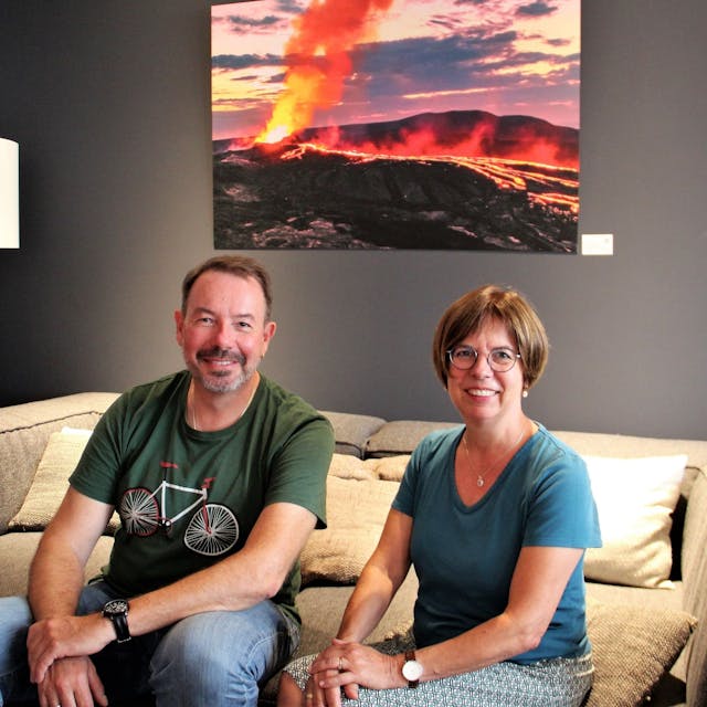 Ein Mann mit Bart und eine Frau mit Pagenkopf und Brille sitzen auf einem Sofa, hinter sich ist ein Bild von einem Vulkan-Ausbruch zu sehen.&nbsp;