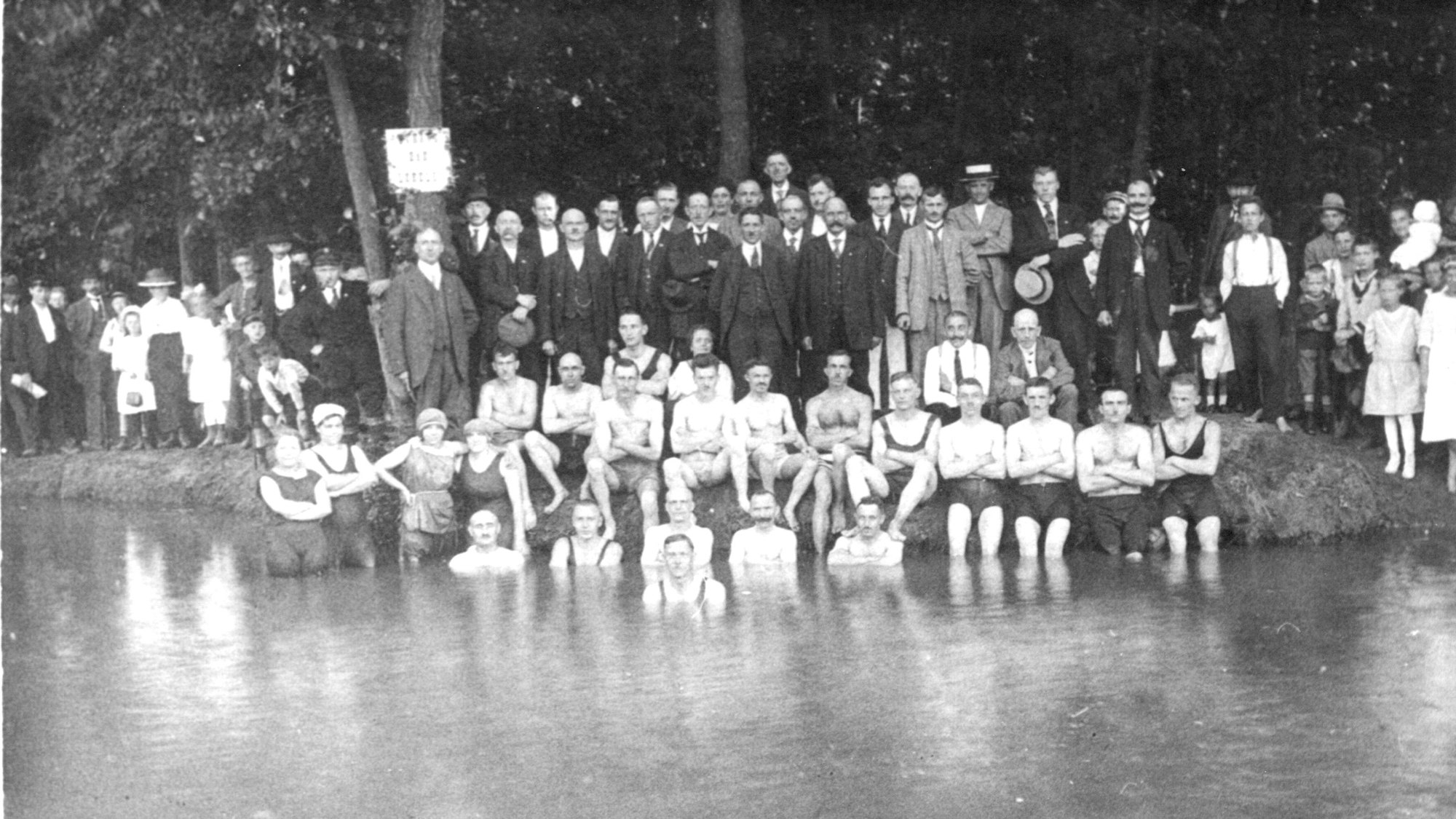 Auf einer Schwarz-Weiß-Fotografie sind viele Männer im Anzu zu sehen. Vor ihnen sitzen junge Leute am Ufer eines Sees, zum Teil im Wasser.