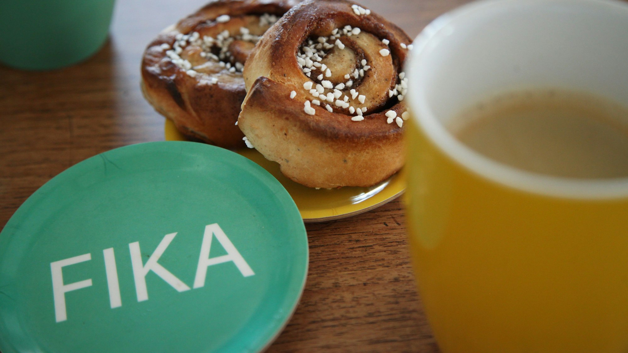 Ein Unterteller mit dem Aufdruck „Fika“ liegt neben zwei Zimtschnecken und zwei Bechern mit Kaffee. Fika bedeuteet „Kaffee trinken“ oder „Kaffeepause machen“.