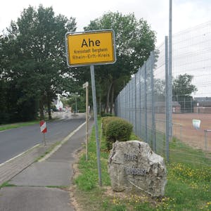 Das Bild zeigt das Ortsschild von Ahe, daneben eine Straße und den Aschenplatz des Dorfs.