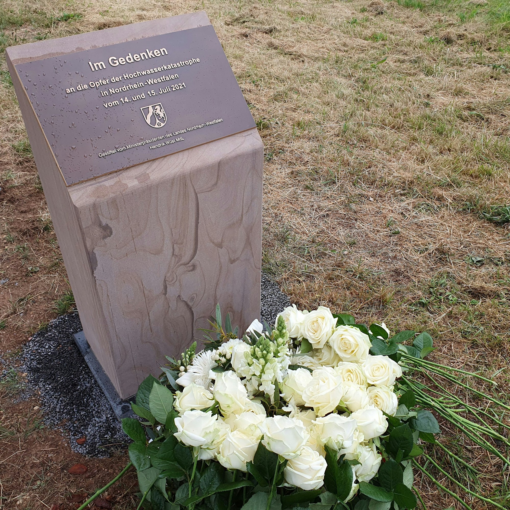 Auf einem Denkmal steht: „In Gedenken an die Opfer der Hochwasserkatastrophe in Nordrhein Westfalen vom 14. und 15. Juli 2021“. Darunter liegen weiße Rosen.