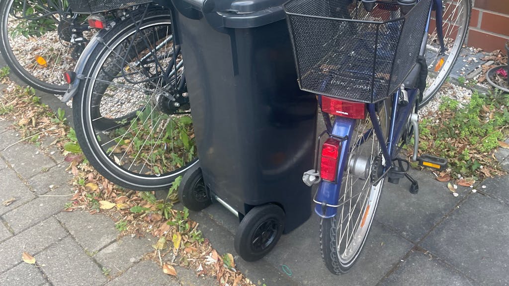 Eine Mülltonne, die zwischen Fahrrädern direkt neben einem Fußweg steht.&nbsp;