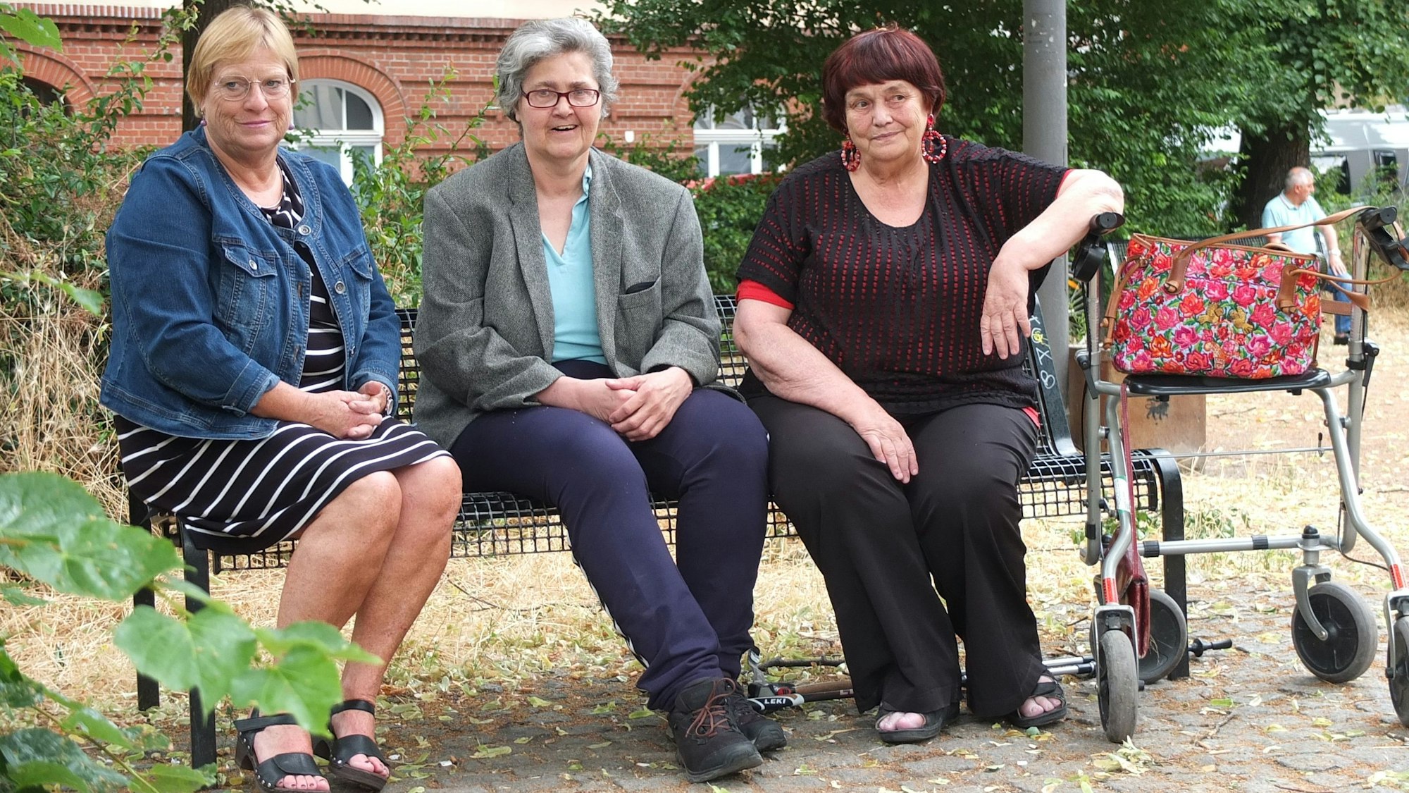 Drei ältere Damen sitzen auf einer Bank, eine stützt den Arm auf ihren Rollator.