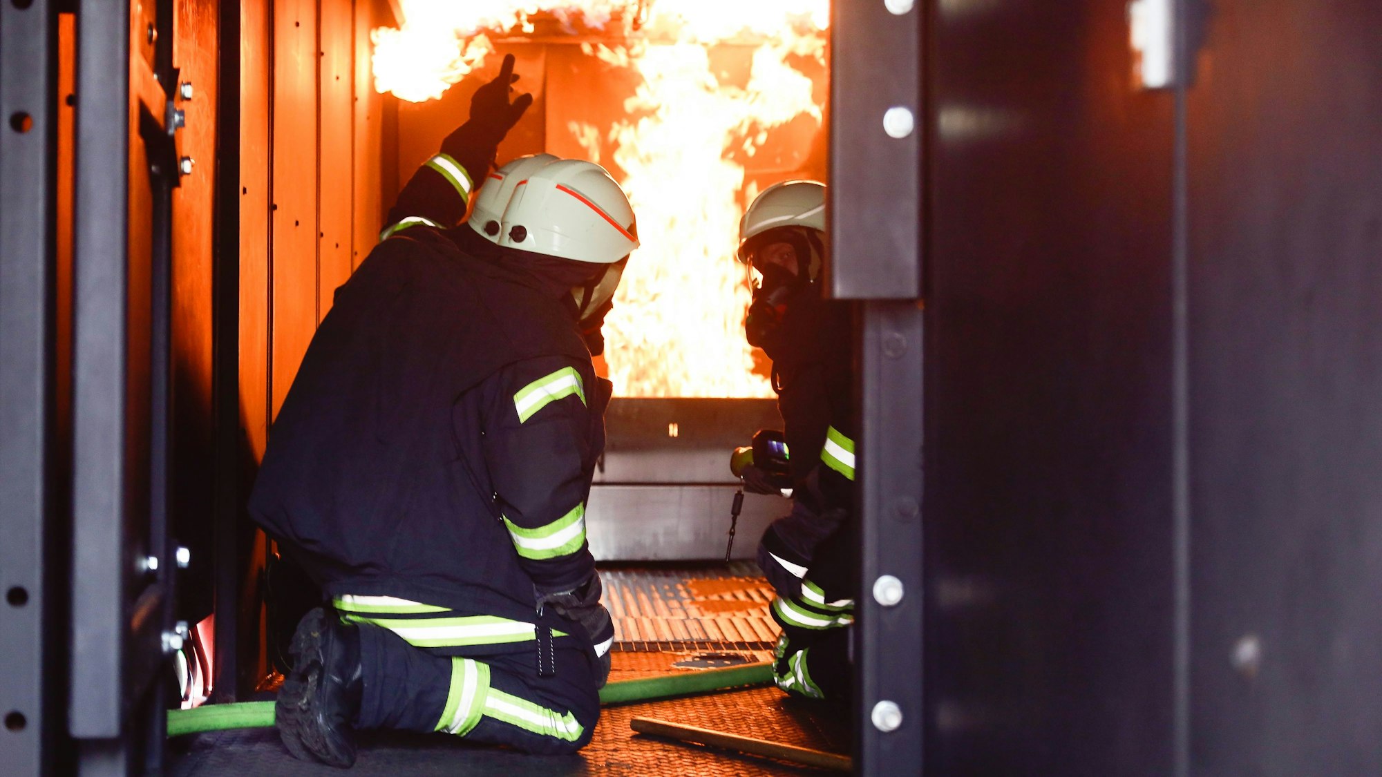 Zwei Feuerwehrmänner knien im Brandübungscontainer der Berufsfeuerwehr Köln vor einer Flammenwand.