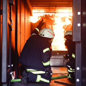 Zwei Männer in Feuerwehruniformen knien im Brandcontainer der Feuerwehr, im Hintergrund sind Flammen zu sehen.