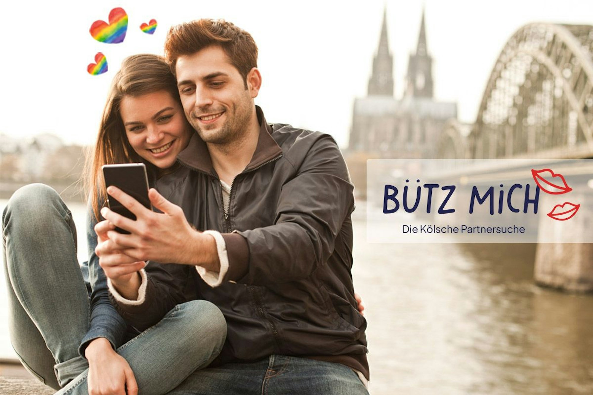 Eine Frau und ein Mann stehen auf der Rheinpromenade mit dem Kölner Dom und der Hohenzollernbrücke im Hintergrund und schauen lächelnd auf ein Smartphone. Daneben das Logo der Website Bütz mich.
