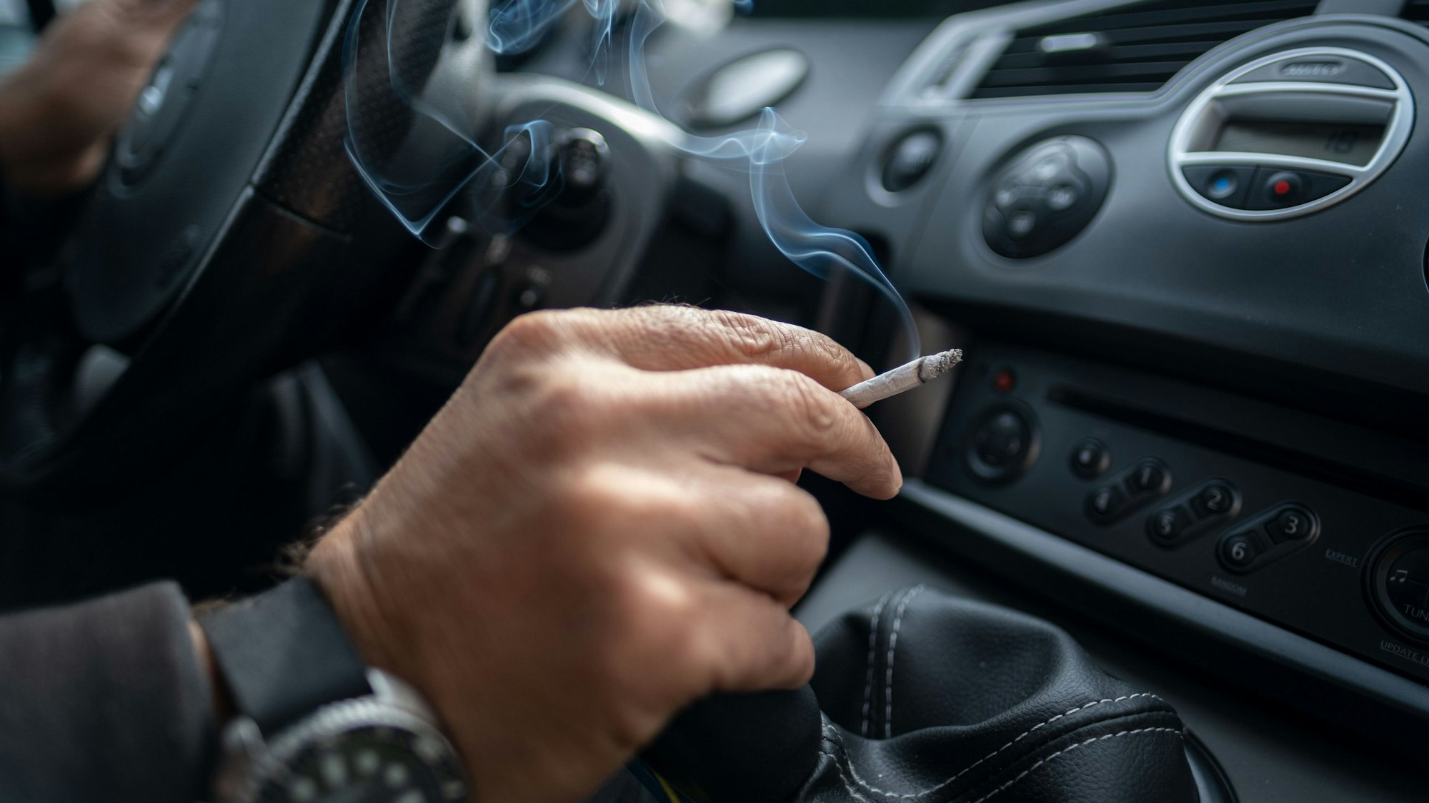 Ein Mann sitzt rauchend am Lenkrad eines Autos. Wer im Auto raucht, schadet allen Mitfahrern. Eine Initiative mehrerer Länder will vor allem Kinder und Jugendliche per Gesetz vor dem Qualm schützen.