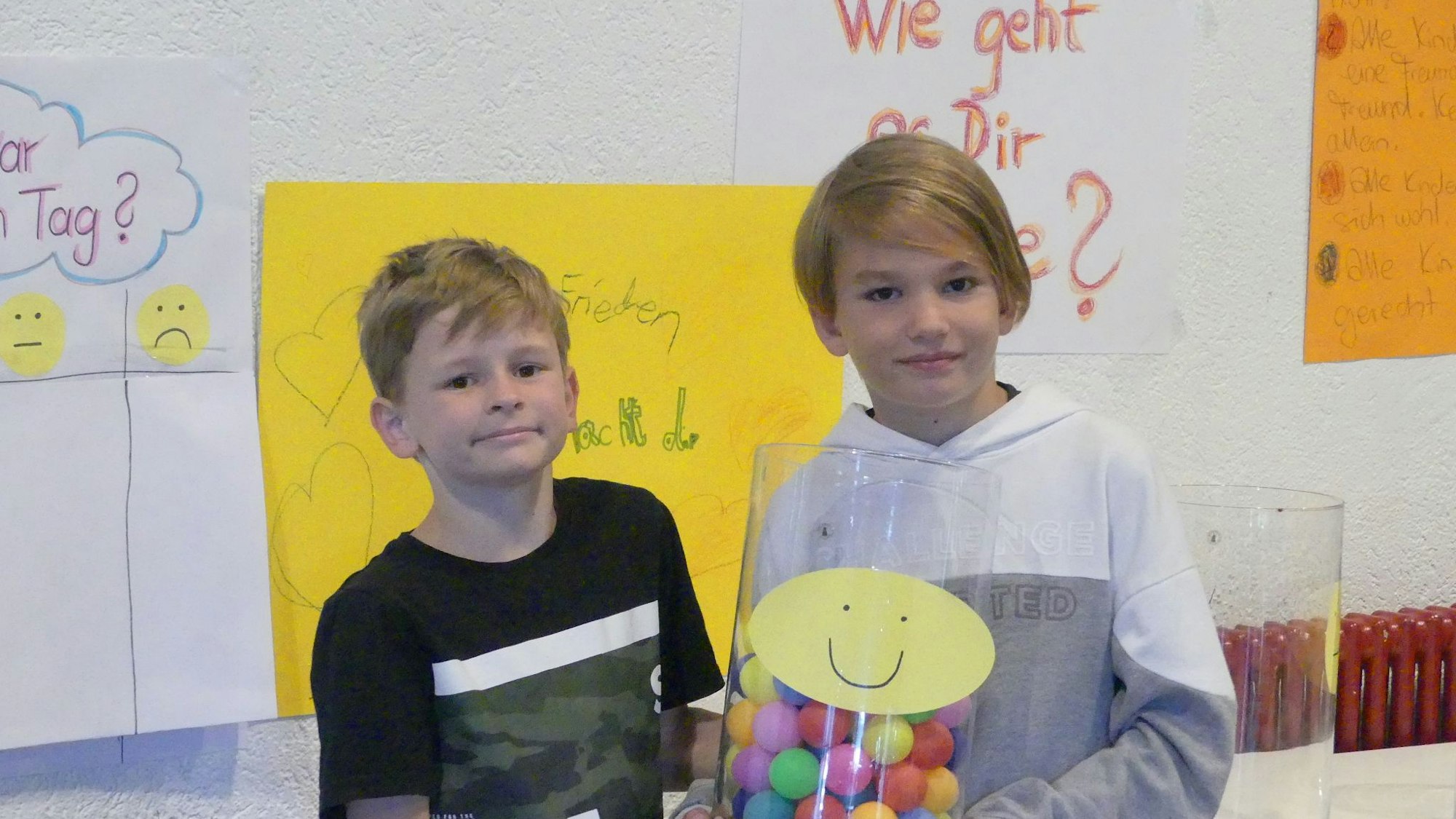 Zwei Kinder stehen vor einem gelben Plakat. Eins hat ein Glas mit bunten Bällen darin in der Hand.
