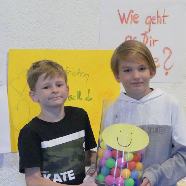 Zwei Kinder stehen vor einem gelben Plakat. Eins hat ein Glas mit bunten Bällen darin in der Hand.