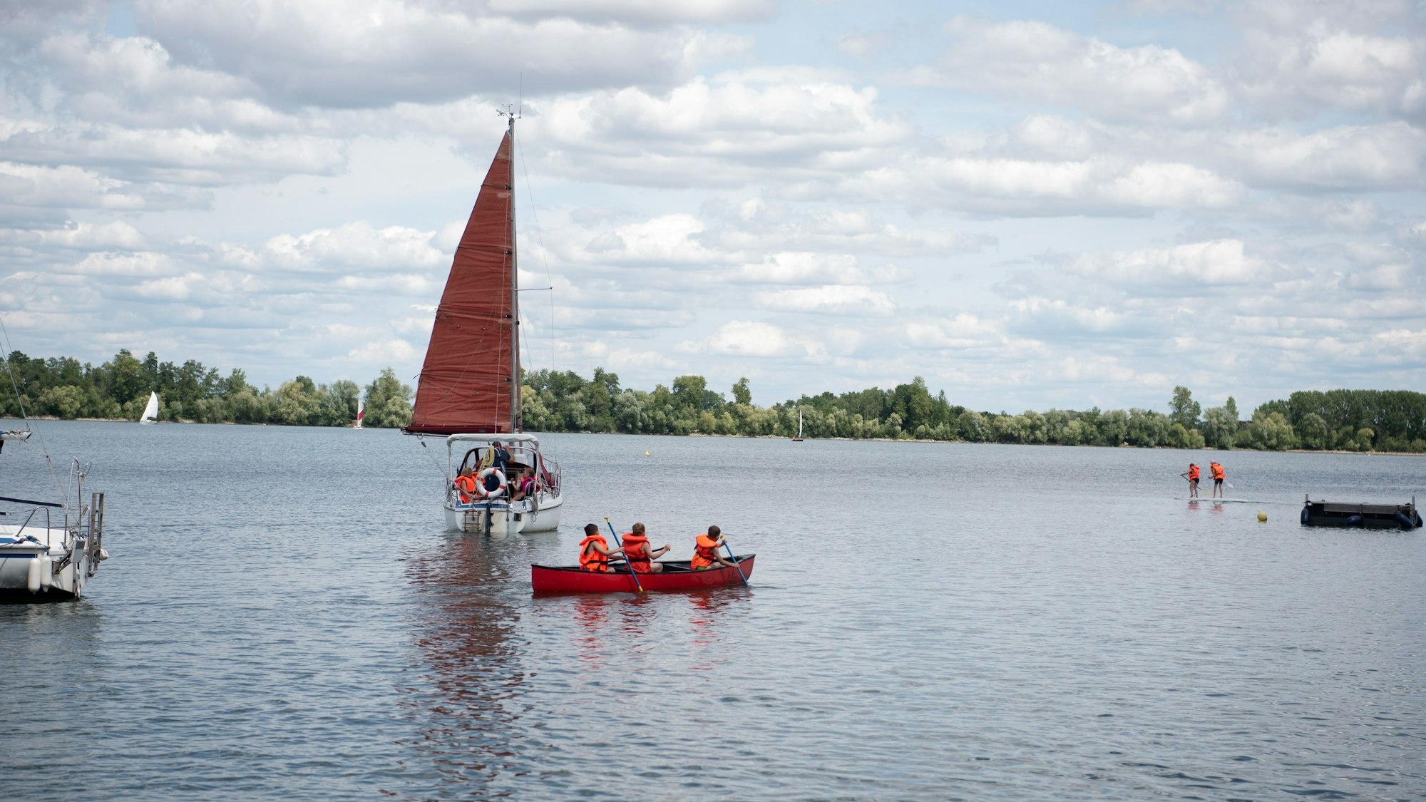 Kinder paddeln in einem Boot auf einem See, dahinter ist ein Segelboot zu sehen.