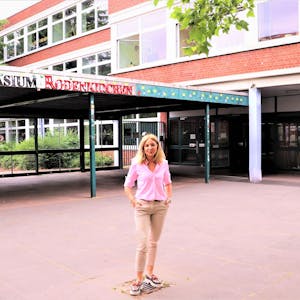 Eine blonde Frau mit schulterlangem blonden Haar steht vor einem Gebäude, auf dem der Schriftzug Gymnasium Rodenkirchen zu lesen ist.