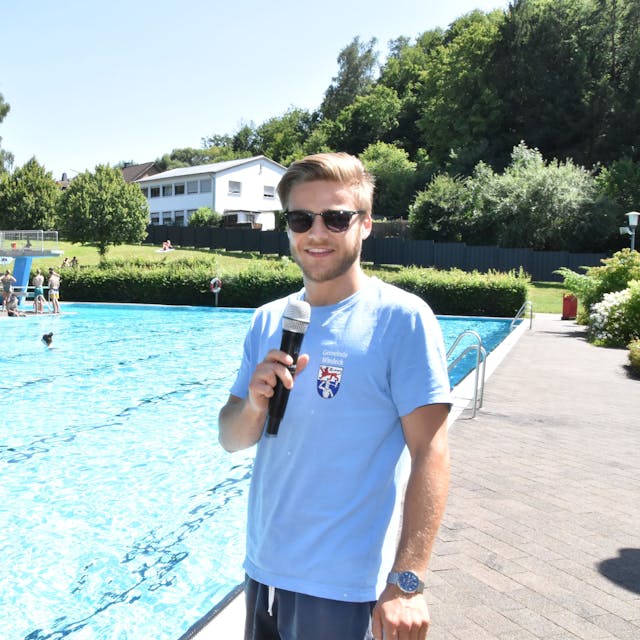 Am Becken eines Freibads steht ein junger Mann mit einem Mikrofon, das ersetzt der Badeaufsicht die Trillerpfeife.