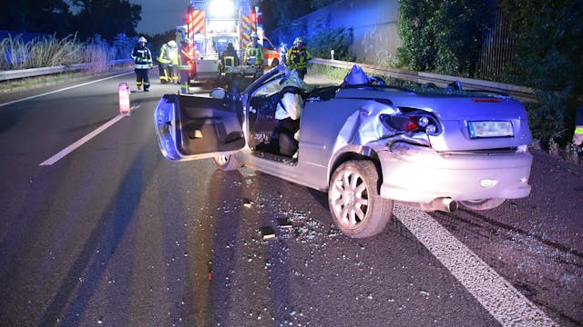 Das zerstörte Fahrzeug steht nach dem Unfall auf der Autobahn A57, im Hintergrund arbeiten Rettungskräfte.