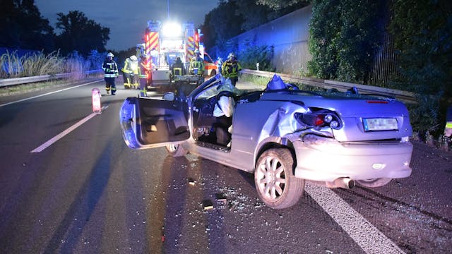 Das zerstörte Fahrzeug steht nach dem Unfall auf der Autobahn A57, im Hintergrund arbeiten Rettungskräfte.