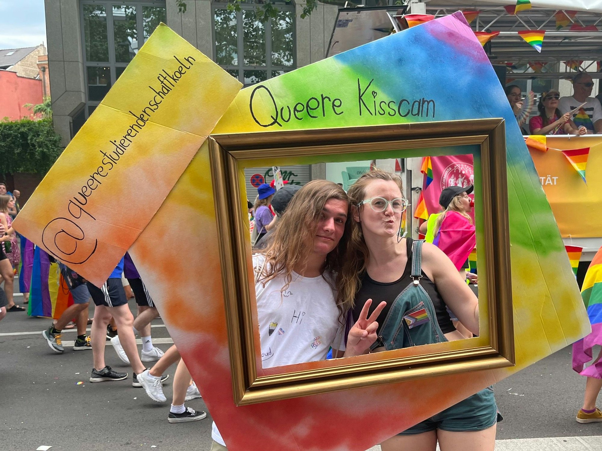Zwei Menschen stehen in einem Bilderrahmen. Zu sehen ist der Schriftzug: Queere Kisscam