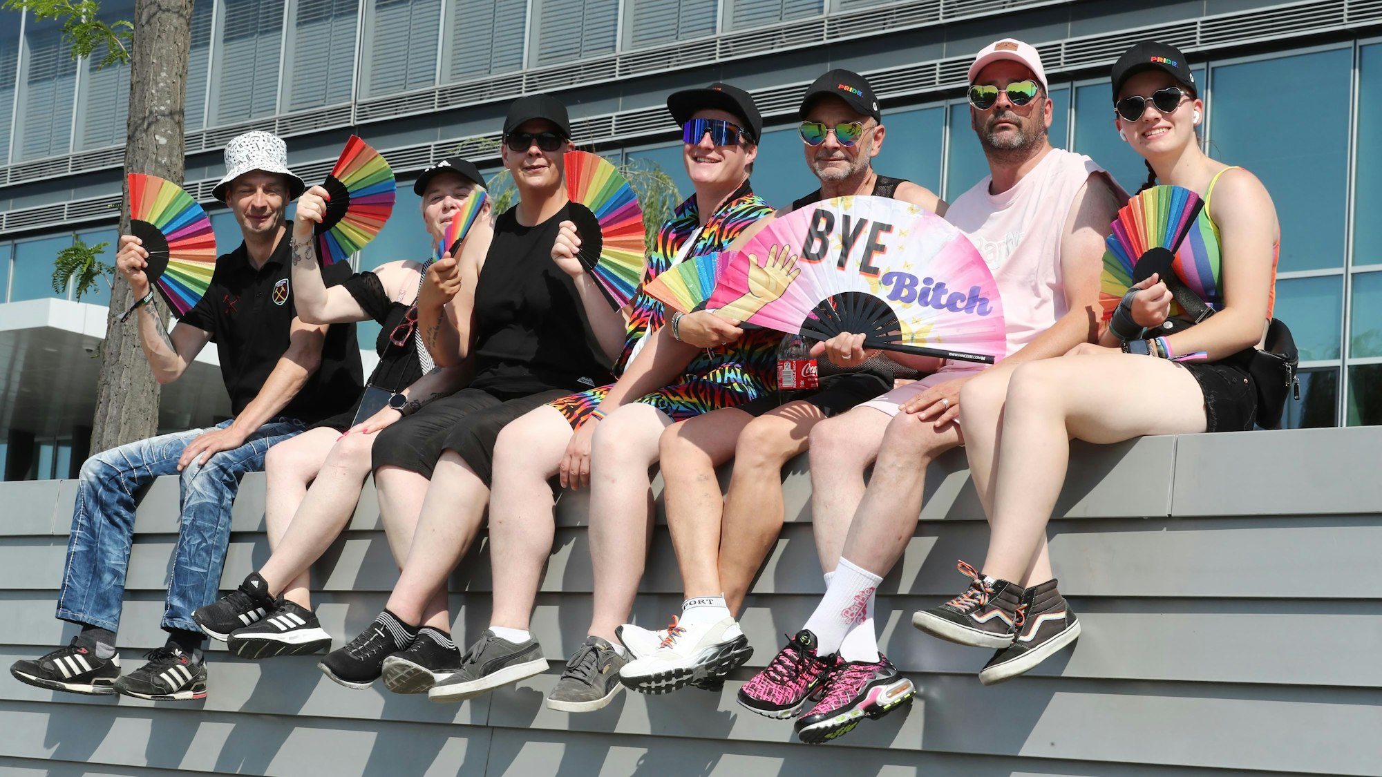 Sieben Teilnehmerinnen und Teilnehmer des Kölner CSD sitzen vor der Mauer eines Gebäudes und halten Fächer in Regenbogenfarben in der Hand.