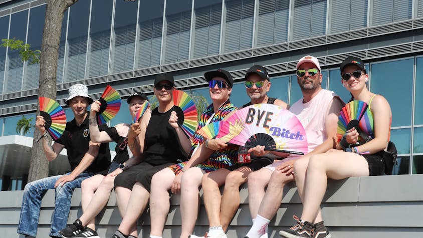 Sieben Teilnehmerinnen und Teilnehmer des Kölner CSD sitzen vor der Mauer eines Gebäudes und halten Fächer in Regenbogenfarben in der Hand.