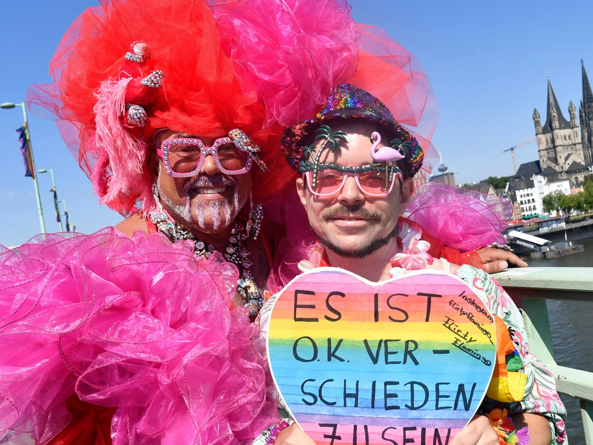 Zwei Menschen stehen in pinker Kleidung vor der Kamera und halten ihr Schild hoch. Im Hintergrund ist der Kölner Dom und Rhein zu sehen.
