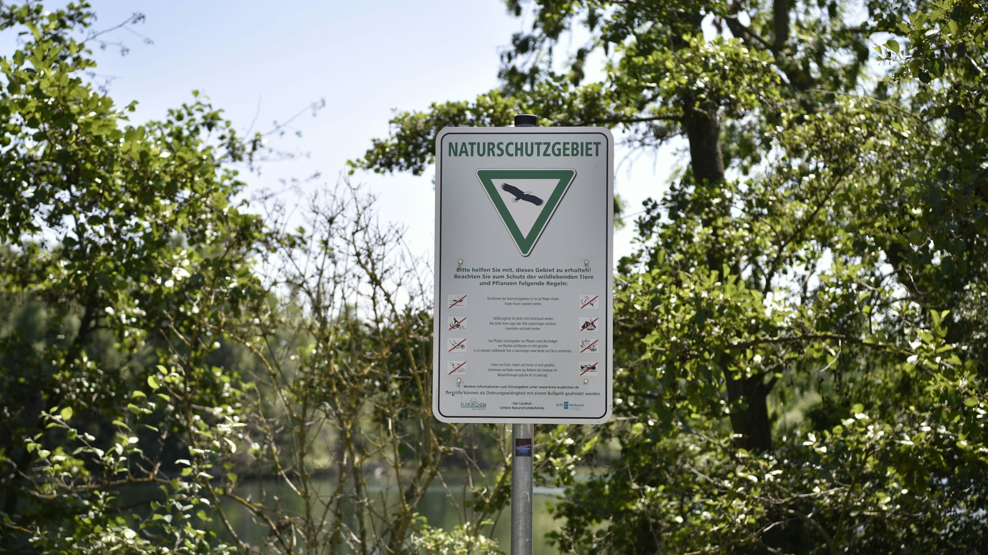 Ein Schild steht vor ein paar Bäumen, im Hintergrund ist Wasser zu sehen. Auf dem Schild steht „Naturschutzgebiet“. Darunter ist das Bild von einem Vogel in einem grünen Dreieck. Darunter wiederum ist eine Auflistung der geltenden Verhaltensregeln zu lesen.