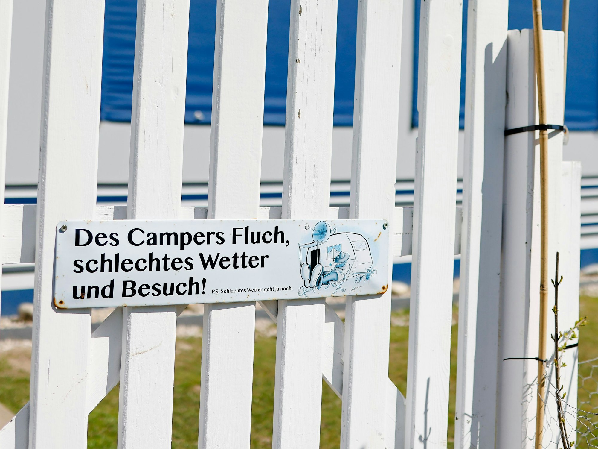 Ein Schild mit dem Spruch "Des Campers Fluch, schlechtes Wetter und Besuch" ist an einem Zaun vor einem Vorzelt auf einem Campingplatz befestigt.