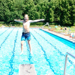 Ein Junge springt rückwärts vom Drei-Meter-Brett, im Hintergrund ist das Schwimmbecken zu sehen.