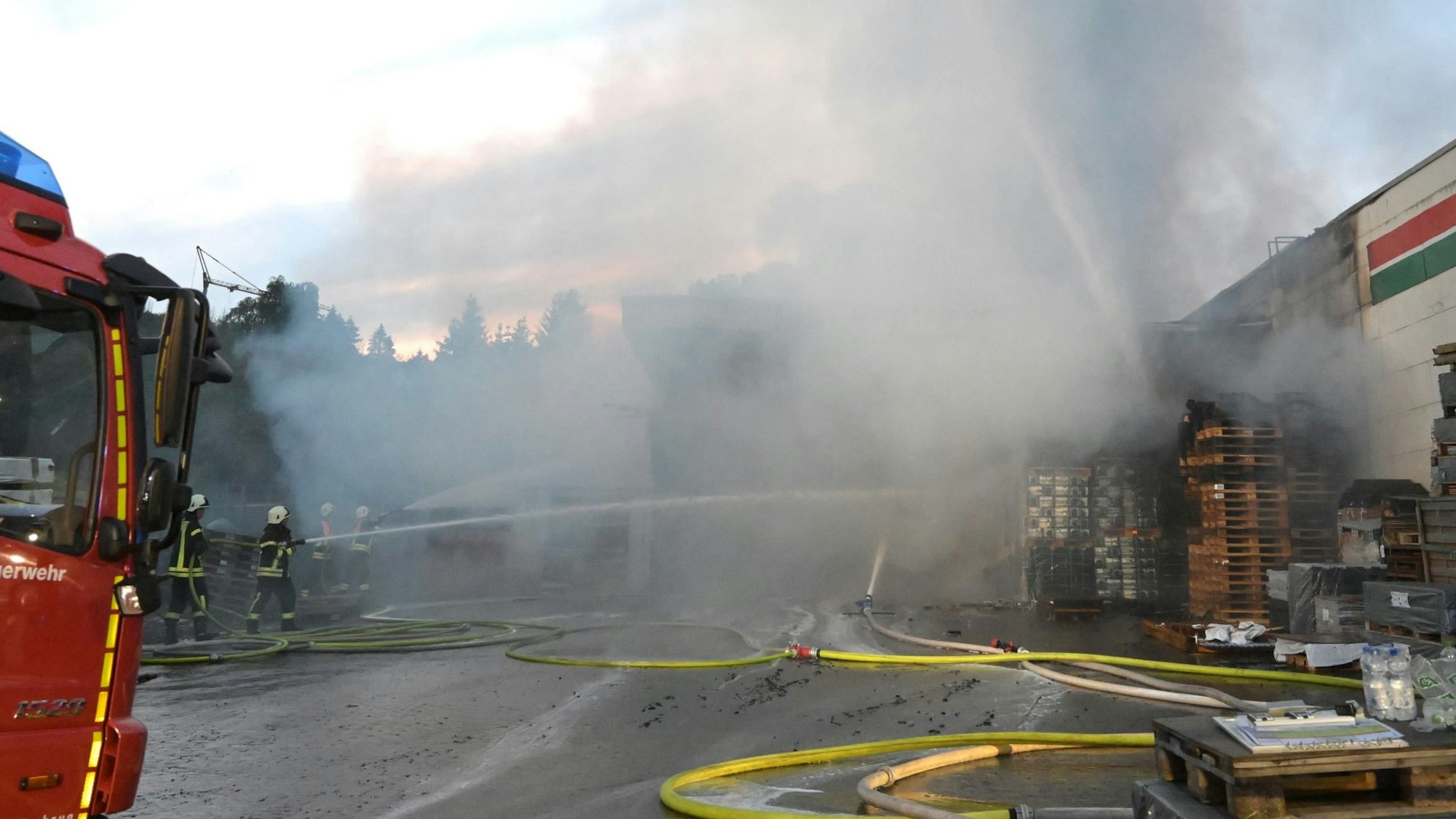 Ein Feuerwehrwagen steht vor einem brennenden Gebäude, auf der ausfahrbaren Leiter stehen Feuerwehrleute und kämpfen mit einem dicken Wasserstrahl gegen die Flammen.