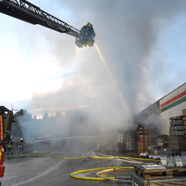 Ein Feuerwehrwagen steht vor einem brennenden Gebäude, auf der ausfahrbaren Leiter stehen Feuerwehrleute und kämpfen mit einem dicken Wasserstrahl gegen die Flammen.&nbsp;
