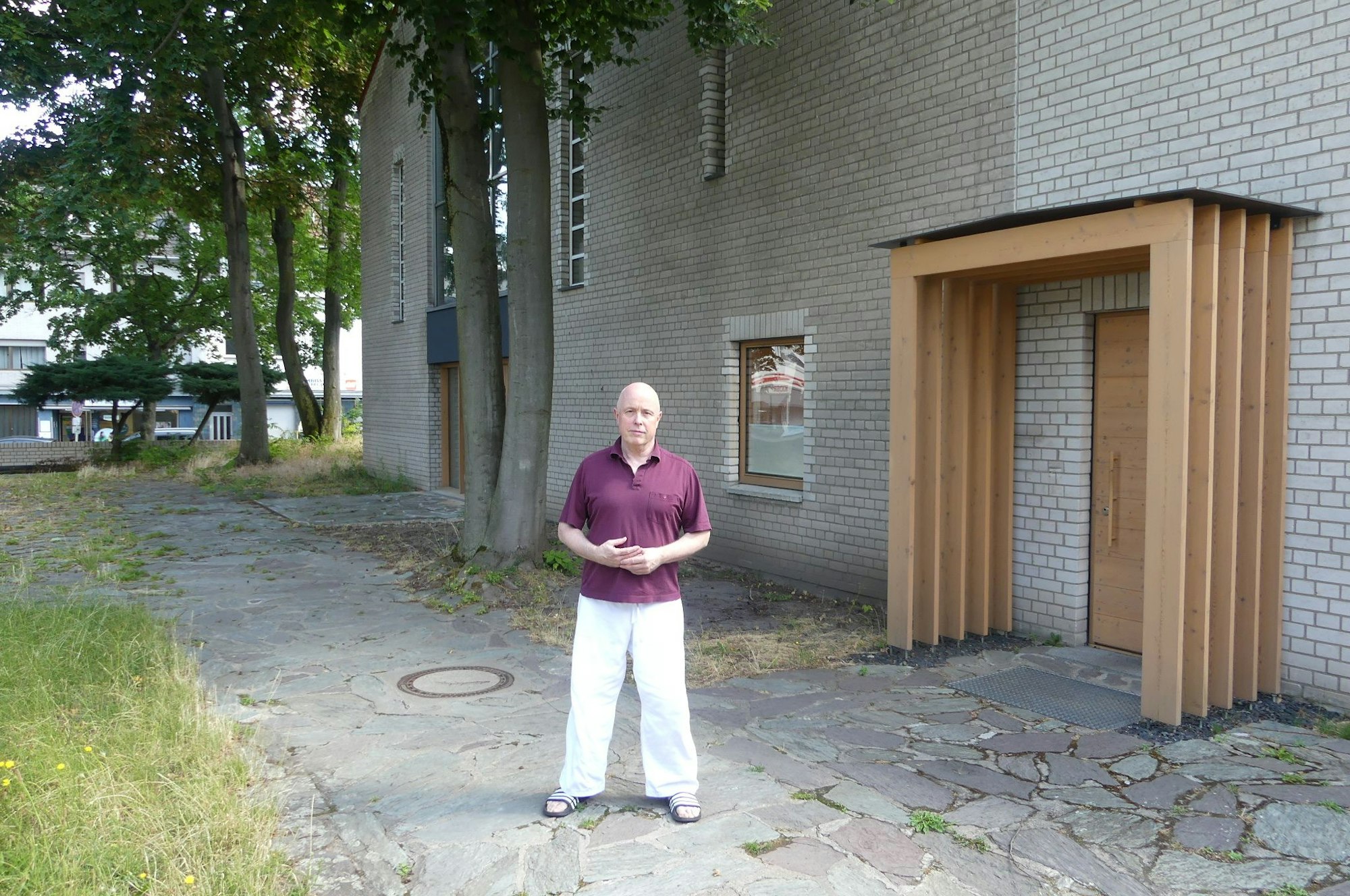 Ein älterer Mann im weinroten Shirt und weißer Hose steht vor der Kirche.