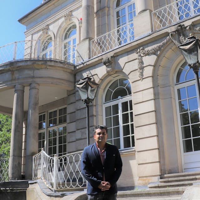 Ein Mann mit dunklem Haar und blauem Jackett steht vor einer großen Villa mit Säulen und Terrassentüren mit Rundbögen.