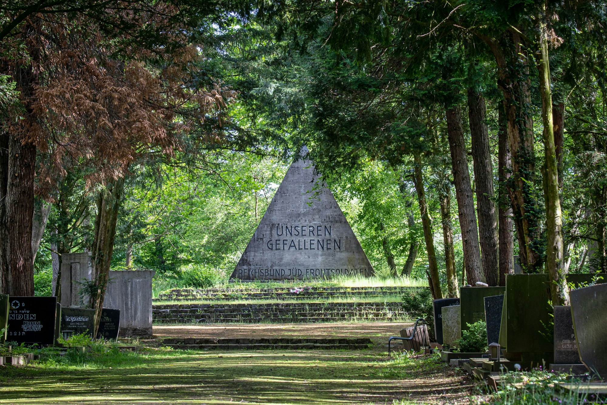 Das Denkmal ist eine graue Pyramide, auf der "unseren Gefallenen" steht und darunter "Reichsbund Jüd. Frontsoldaten". Es ist umgeben von Bäumen.