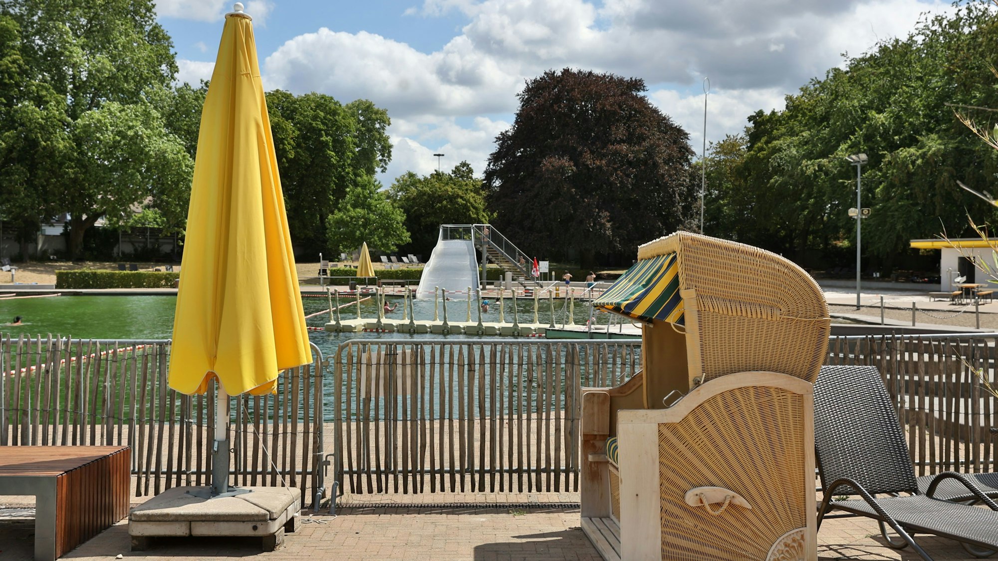 Das Bild zeigt einen Strandkorb und einen geschlossenen, gelben Sonnenschirm um Lentpark Freibad in Köln. Im Hintergrund sind die Rutsche uns das Naturbecken zu sehen,