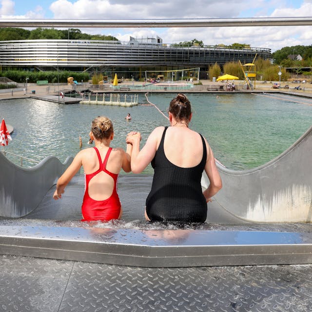 Das Bild zeigt eine Mutter und ihre Tochter von hinten, die einen Wasserrutsche hinunterrutschen. Im Hintergrund sieht man den Naturbadeteich im Lentpark Freibad in Köln.